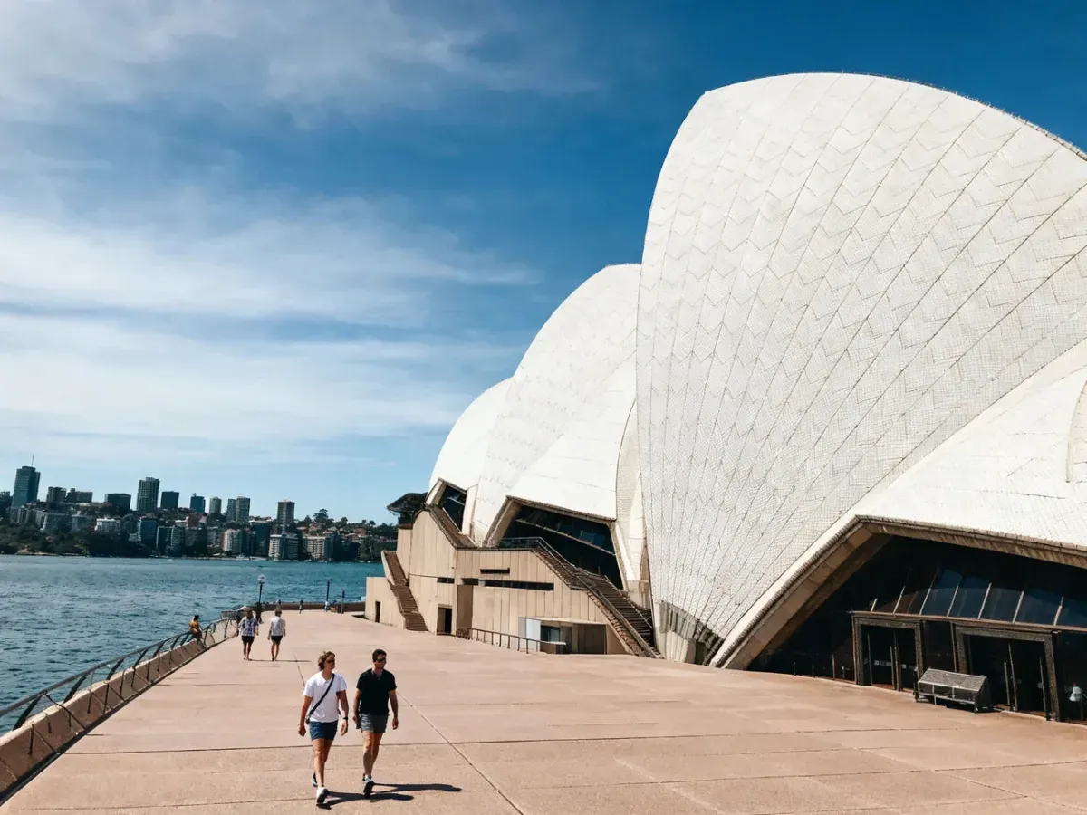 Opera w Sydney w słoneczny dzień, białe żagle nad zatoką, turyści spacerują, zachęcający klimat zwiedzania